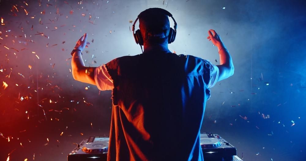 DJ with headphones, hands raised, silhouetted against red and blue lights.