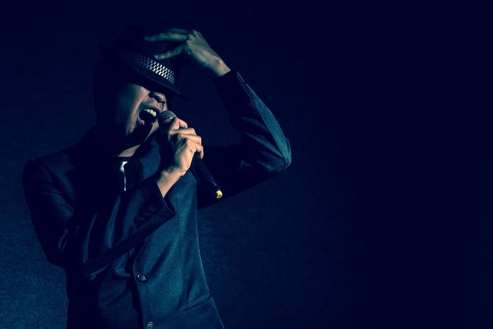 Singer on stage, holding microphone, dark suit, adjusting hat, singing with open mouth, blue lighting.