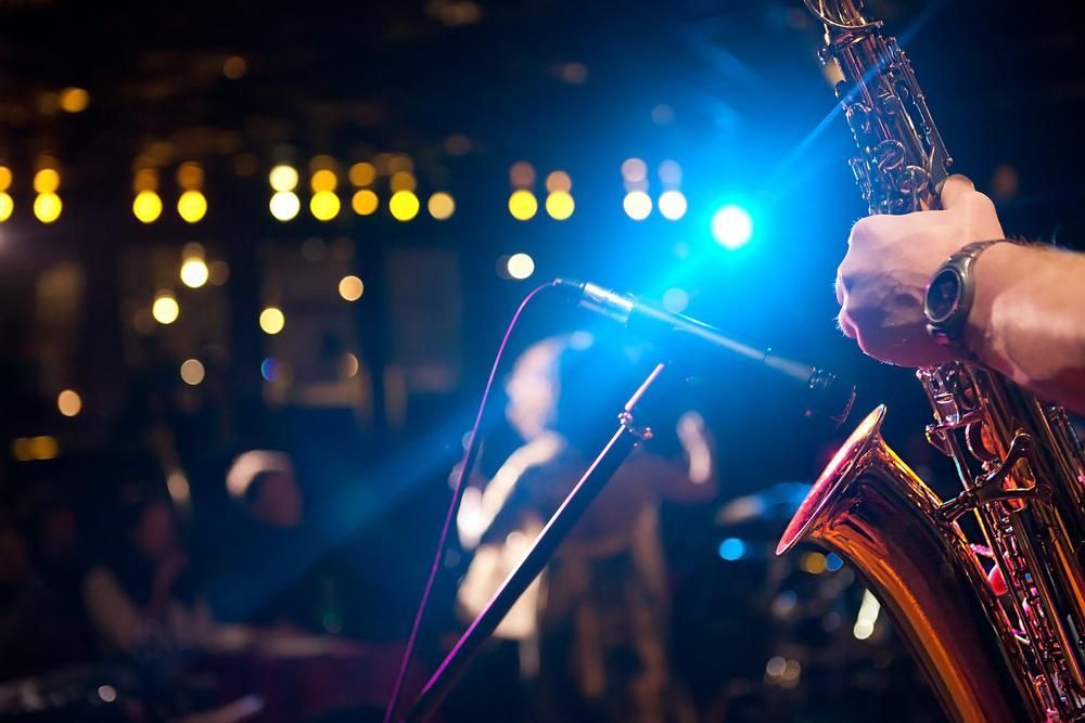 Saxophonist on stage, holding instrument. Blue spotlight, microphone, blurred crowd in background.