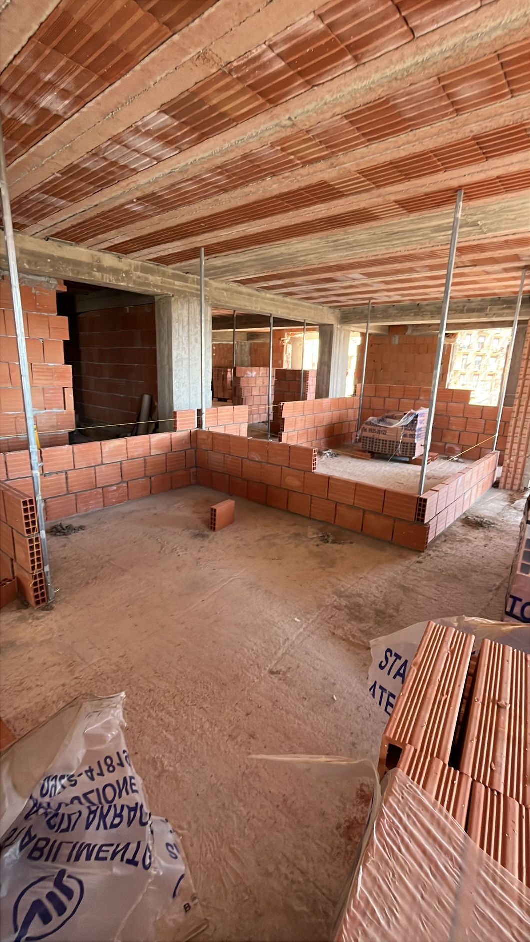 Cantiere edile con muri in mattoni in fase di assemblaggio e travi del soffitto a vista in un'ampia stanza non rifinita.