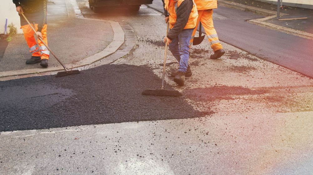 Gli operai stradali, vestiti con indumenti arancioni ad alta visibilità, usano scope a manico lungo per stendere asfalto nero fresco su una strada.