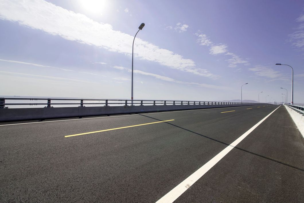 Sotto un cielo azzurro brillante con leggere nuvole, un ponte asfaltato si estende verso l'orizzonte, fiancheggiato da lampioni.