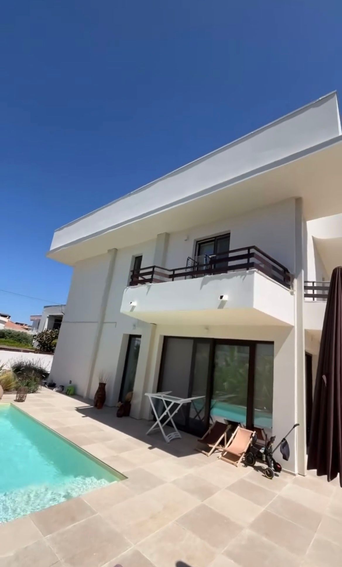 Una casa bianca a due piani con balcone e porte scorrevoli in vetro accanto a una piscina rettangolare sotto un cielo azzurro.