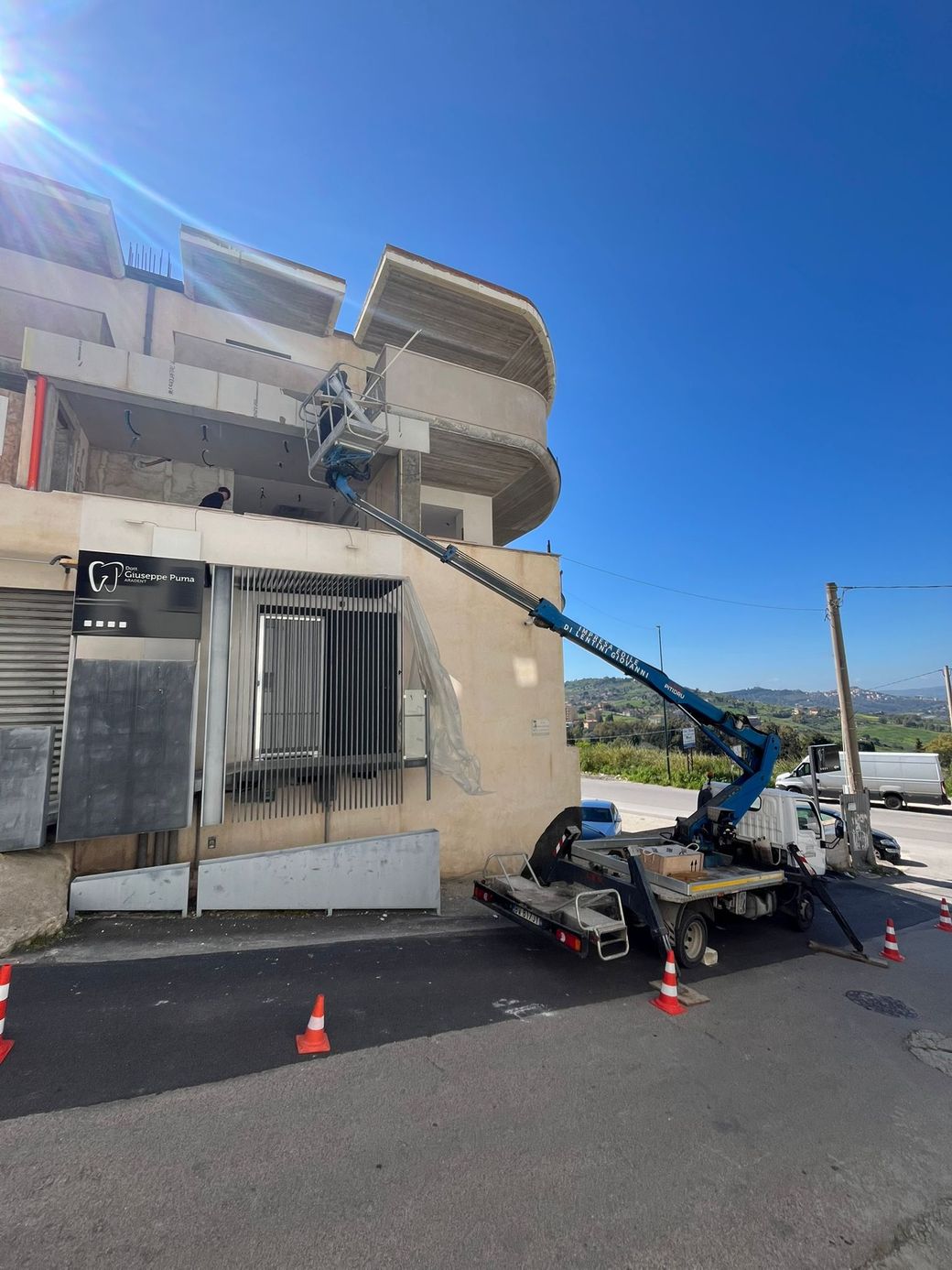 Un carrello elevatore idraulico blu, parcheggiato in strada, si protende verso l'alto per demolire l'angolo di un edificio beige danneggiato.