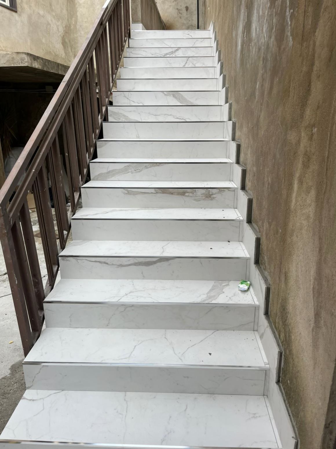 Una ripida scalinata con piastrelle che riproducono il marmo bianco, una ringhiera in legno sulla sinistra e una semplice parete in cemento sulla destra.