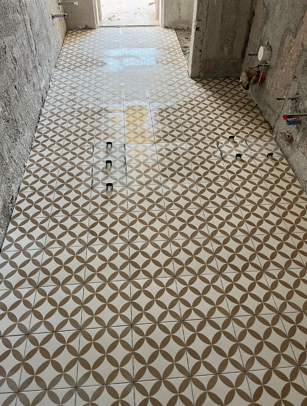 Il pavimento di un corridoio, attualmente in fase di ristrutturazione, è piastrellato con un motivo geometrico ripetuto color crema e marrone, lasciando le pareti a vista.