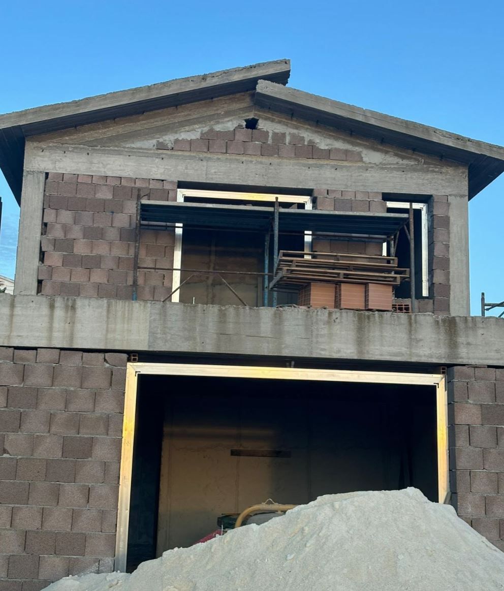 Un edificio a due piani in costruzione con muri in mattoni marroni a vista, struttura in cemento armato e un grande cumulo di sabbia davanti.