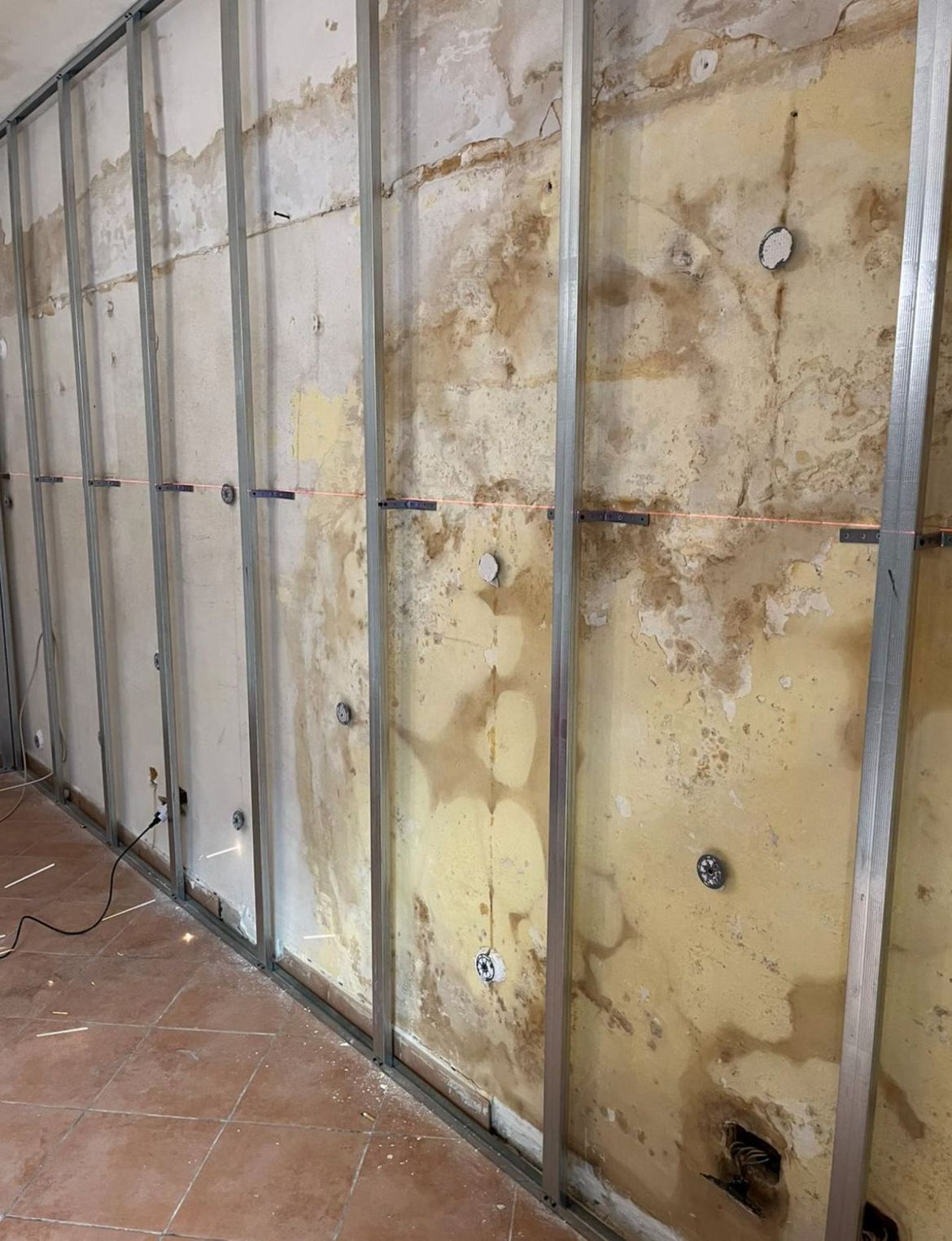 Montanti metallici verticali installati davanti a una vecchia parete in intonaco macchiata e danneggiata all'interno di una stanza con pavimento piastrellato.