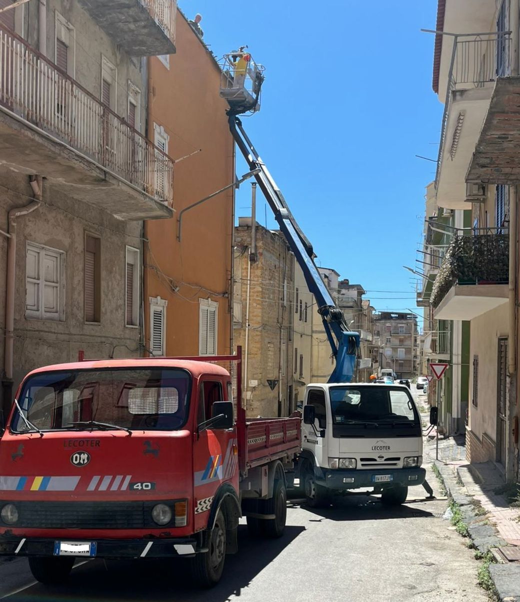 Un camion rosso e un camion bianco parcheggiati su una strada stretta, con una gru a braccio telescopico estesa verso un edificio arancione.