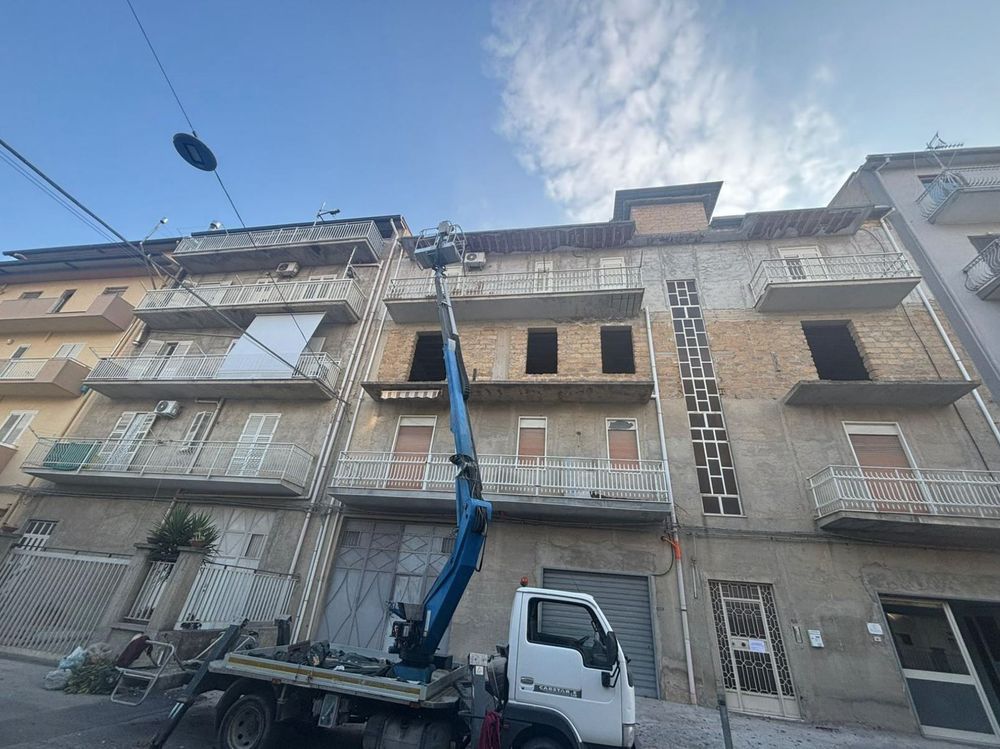 Un camion bianco con una piattaforma aerea blu rialzata è parcheggiato di fronte a un condominio in città, sotto un cielo azzurro.