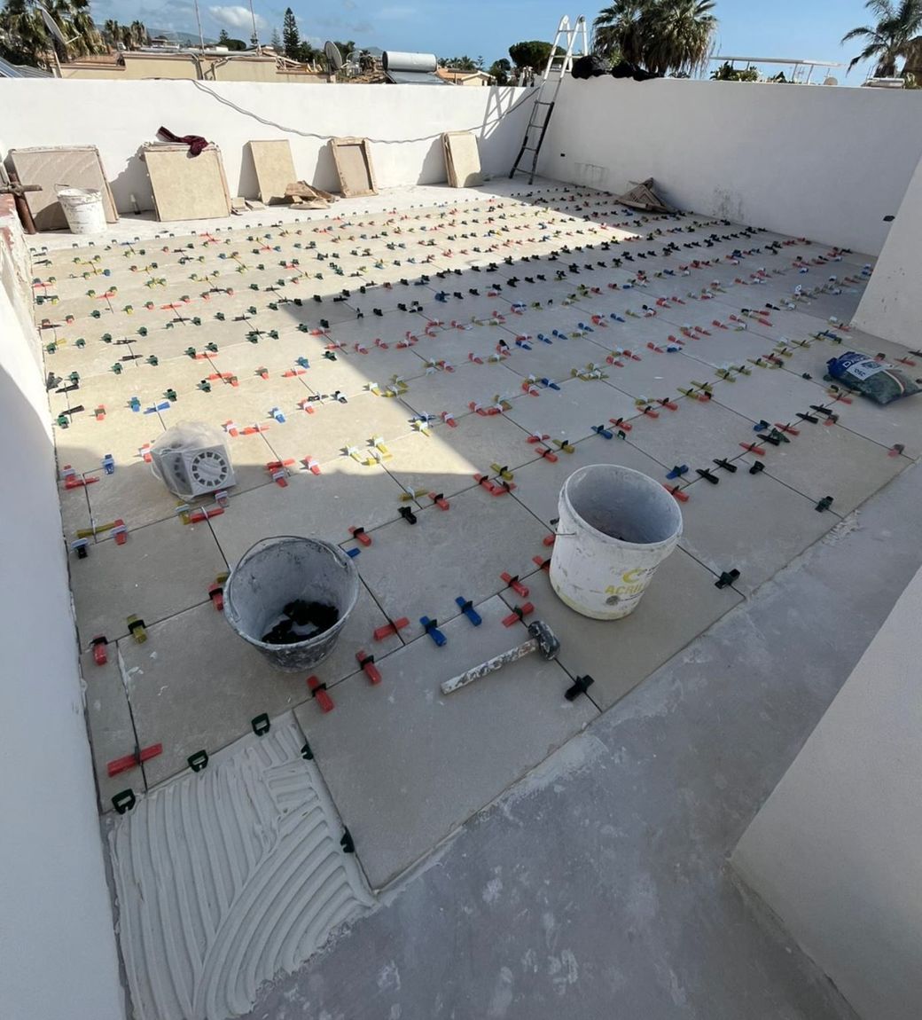 Un pavimento di tetto non rifinito, ricoperto di piastrelle quadrate con distanziatori e due secchi di malta.