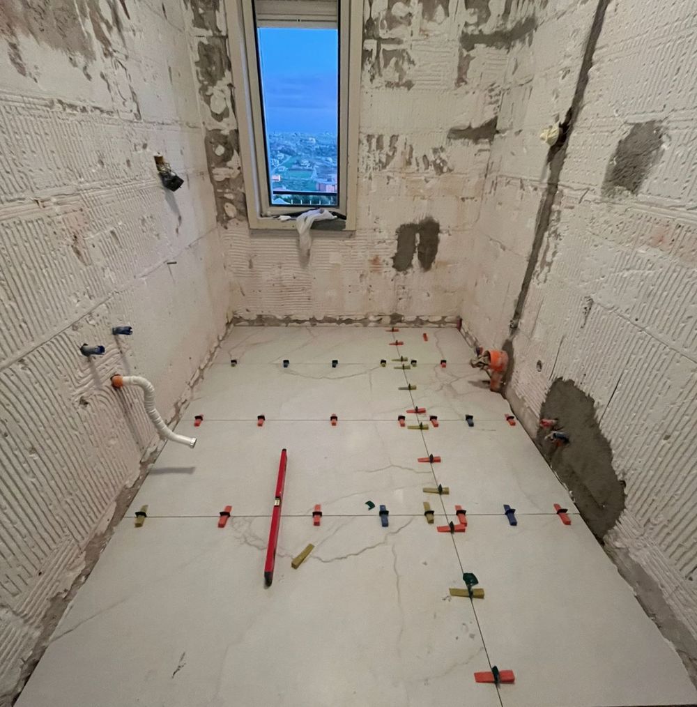 Un bagno in fase di ristrutturazione con piastrelle del pavimento di colore chiaro parzialmente posate, con clip di livellamento e tubature idrauliche a vista.