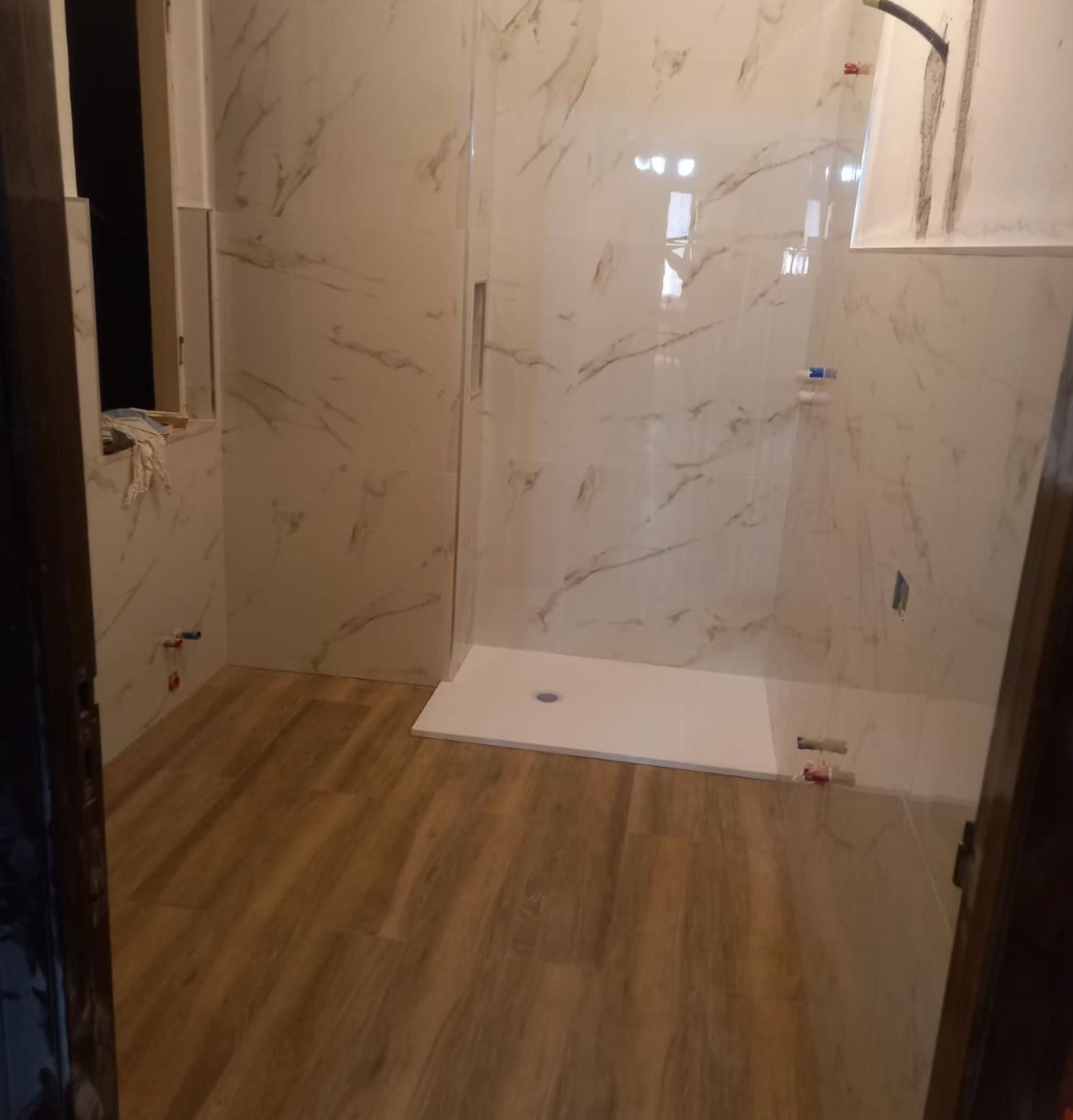 Un bagno moderno con doccia in marmo bianco, pavimento in piastrelle effetto legno e sanitari a parete non rifiniti.