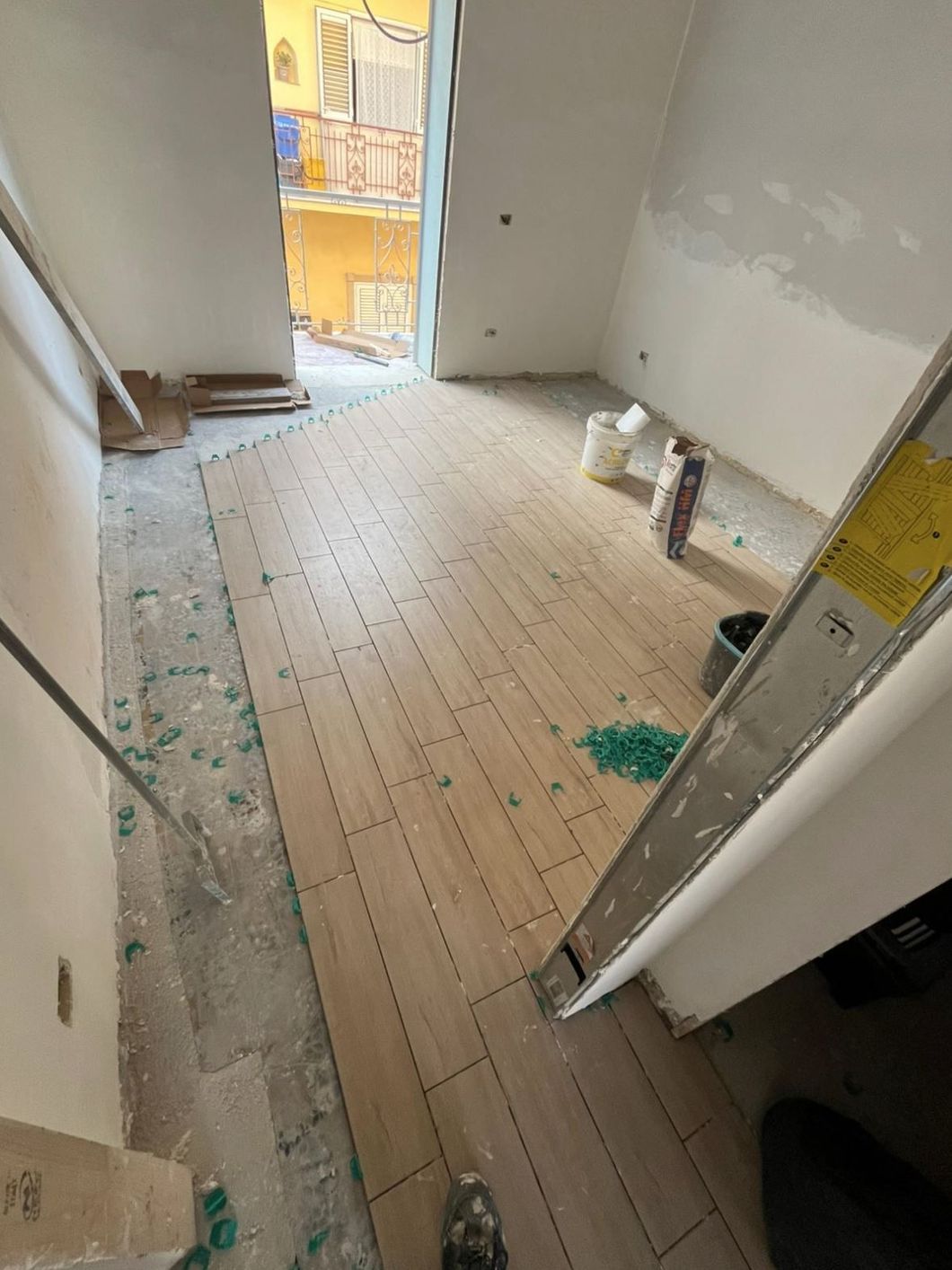 Interno di una stanza in fase di ristrutturazione con pavimento in piastrelle effetto legno chiaro parzialmente posato e distanziatori verdi.