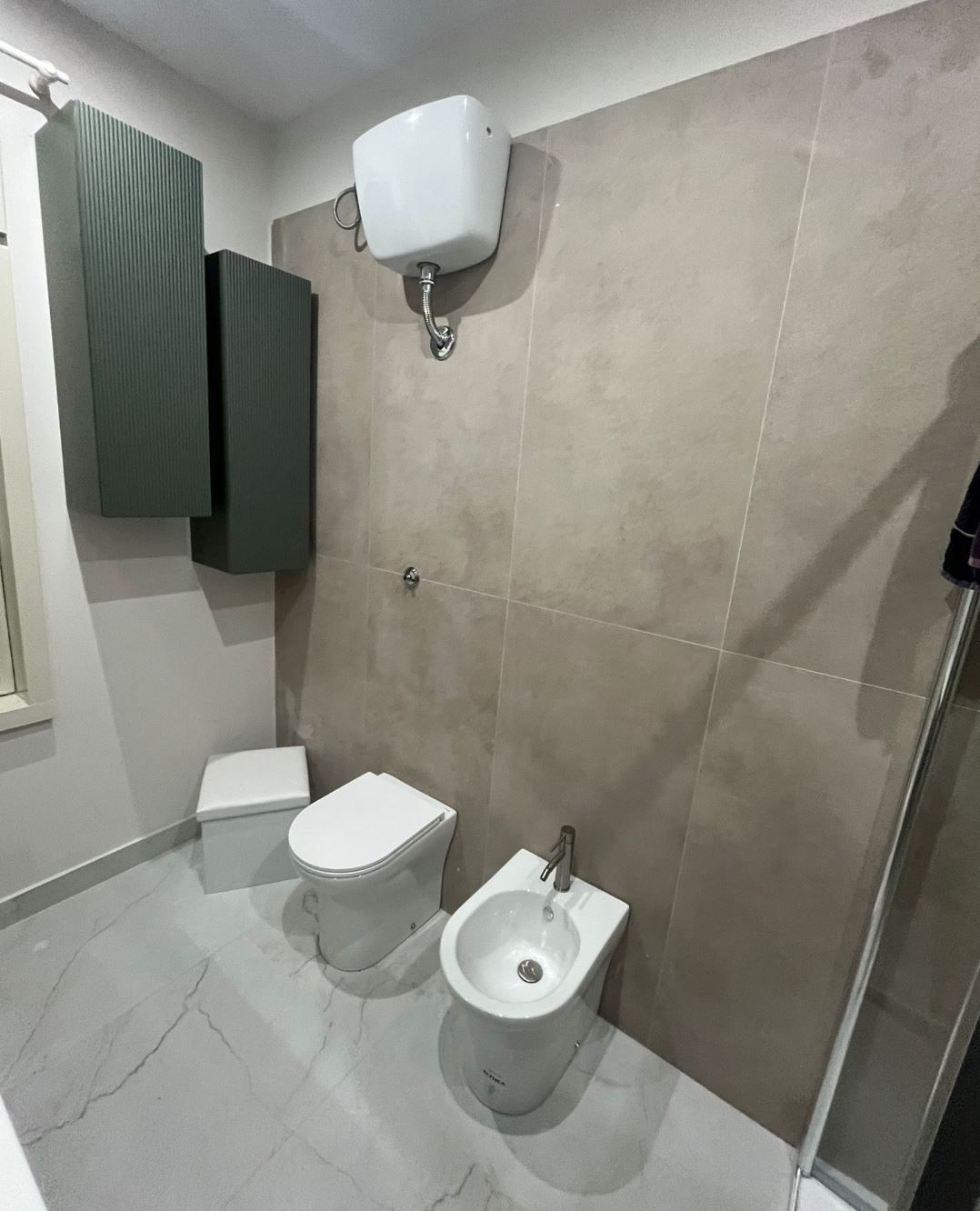 Un bagno piastrellato di beige con WC bianco, bidet, cassetta di scarico a parete e mobili grigio scuro su una parete laterale.