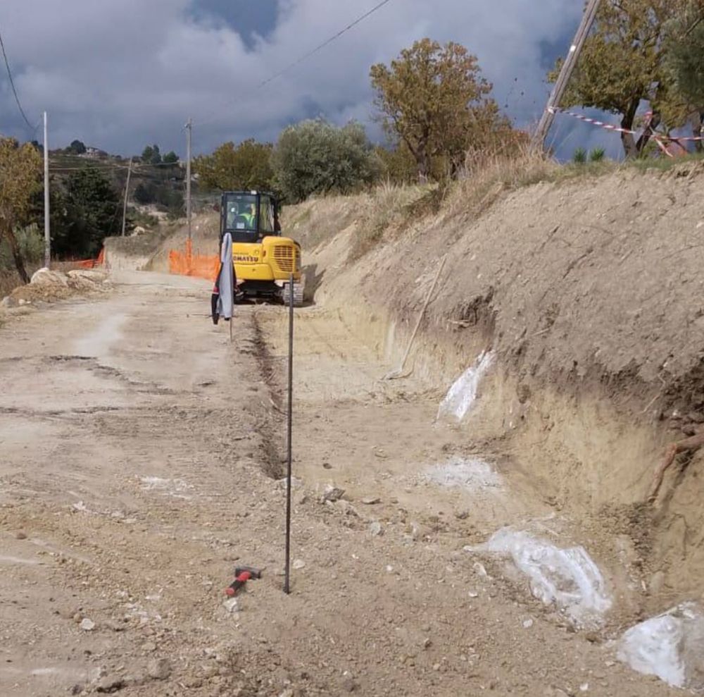 Un miniescavatore giallo esegue lavori di movimento terra lungo una strada sterrata con un ripido terrapieno parzialmente scavato sulla destra.