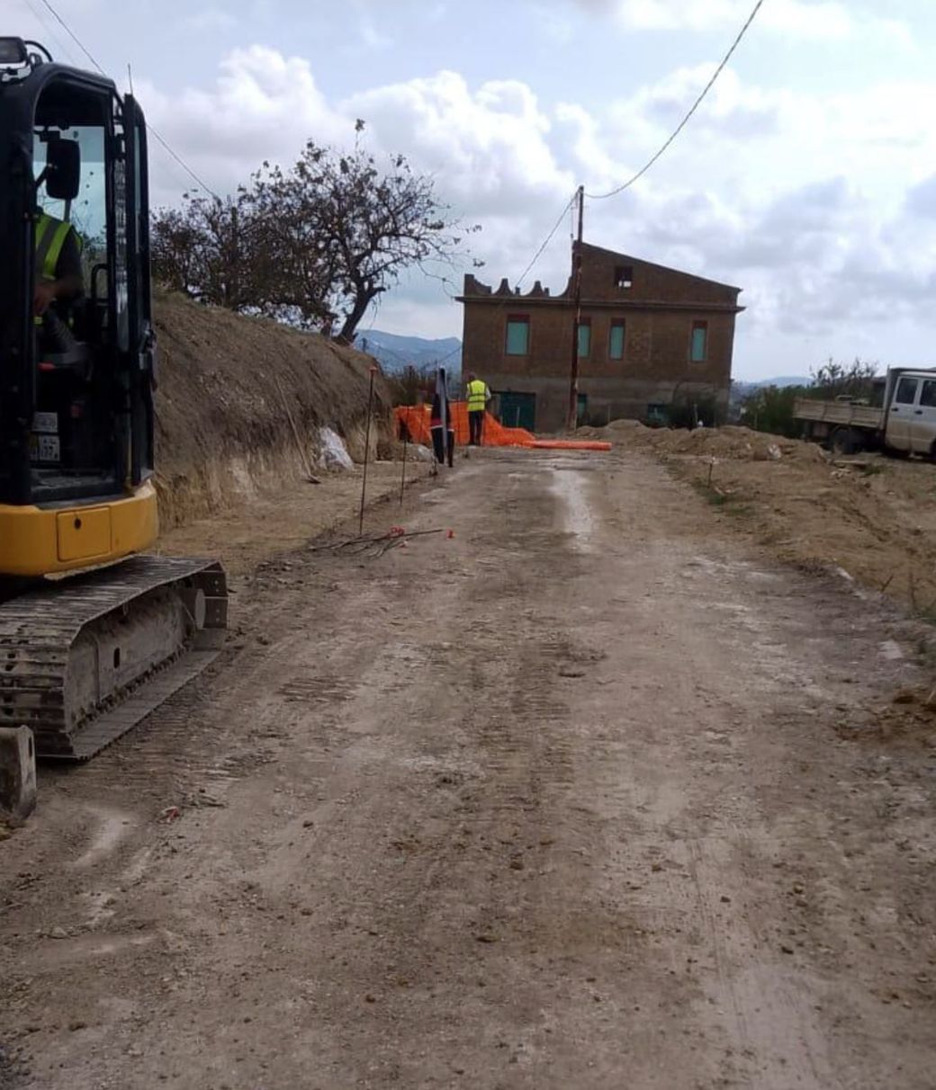 Un escavatore giallo è fermo su una strada sterrata vicino a una barriera di sicurezza arancione e a un edificio in costruzione.