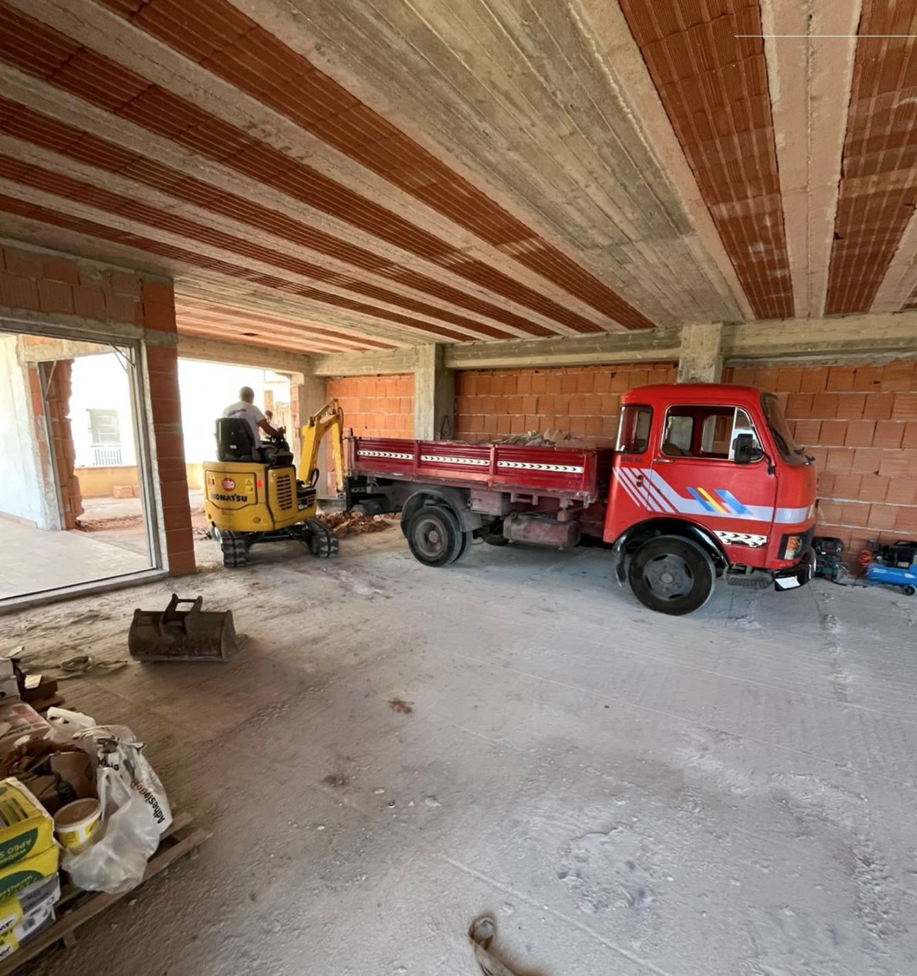 Un escavatore giallo e un autocarro ribaltabile rosso si trovano all'interno di un cantiere edile con muri di mattoni e un soffitto di cemento grezzo.