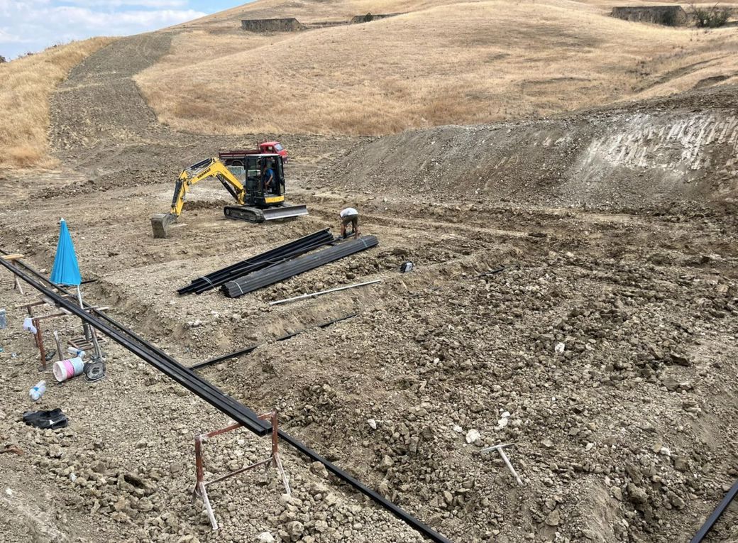 Un escavatore giallo è al lavoro in un cantiere edile su un pendio sterrato, trasportando materiali e attrezzature per lo scavo di trincee.