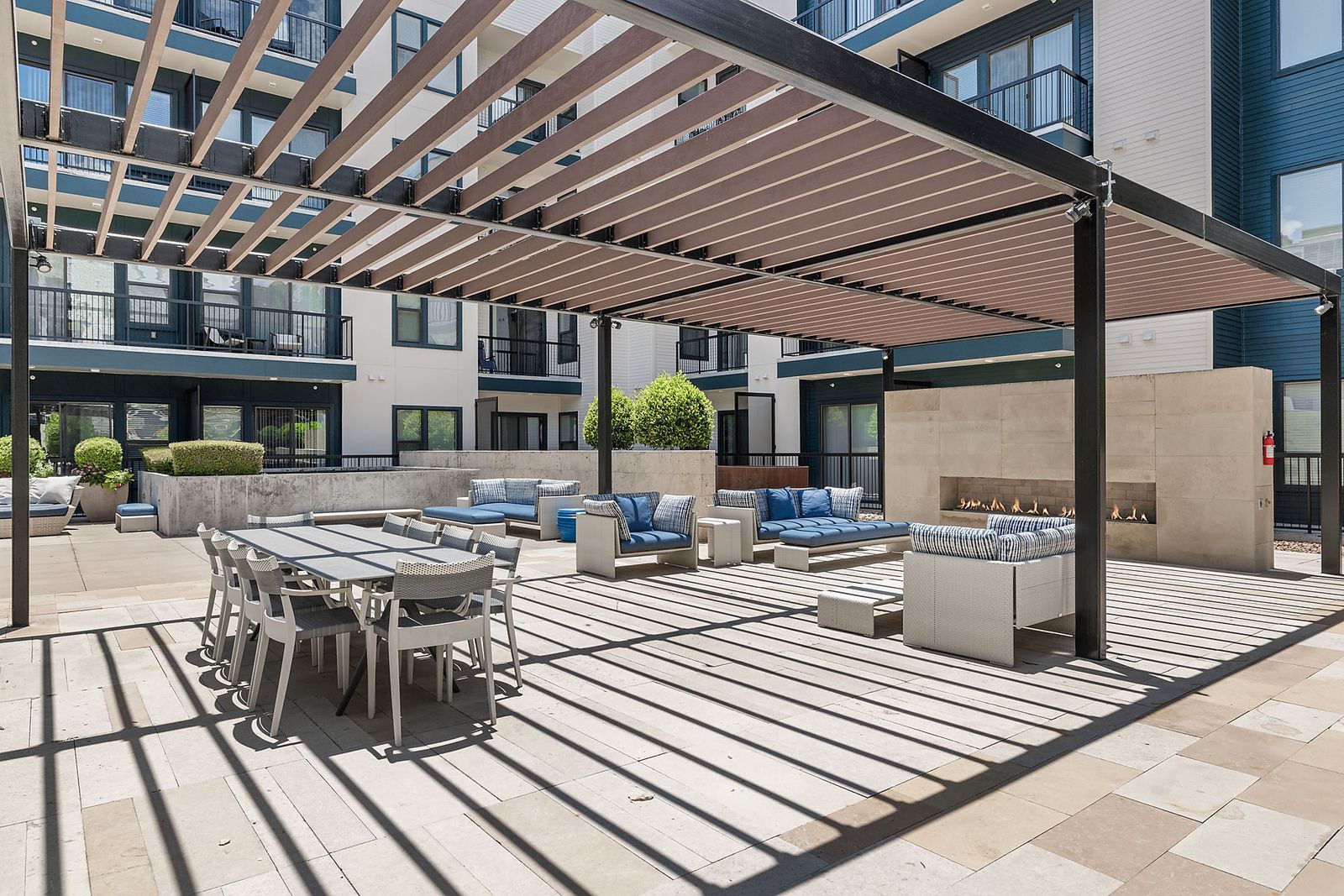 Outdoor courtyard with pergola shade, lounge chairs, dining tables, and modern apartment buildings.