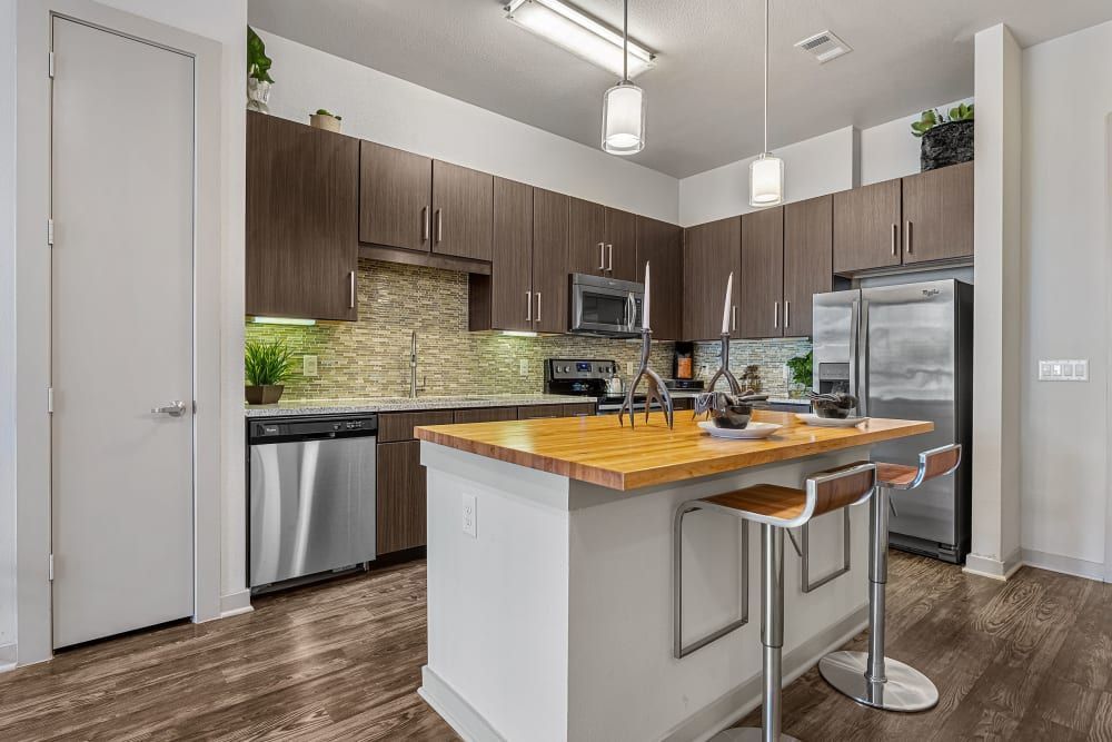 A modern kitchen featuring dark brown wooden cabinetry, stainless steel appliances, a mosaic tile backsplash, and a center island with a butcher block countertop and bar stools. The room is well-lit with pendant lighting and natural tones at Marq Uptown in Austin, TX.