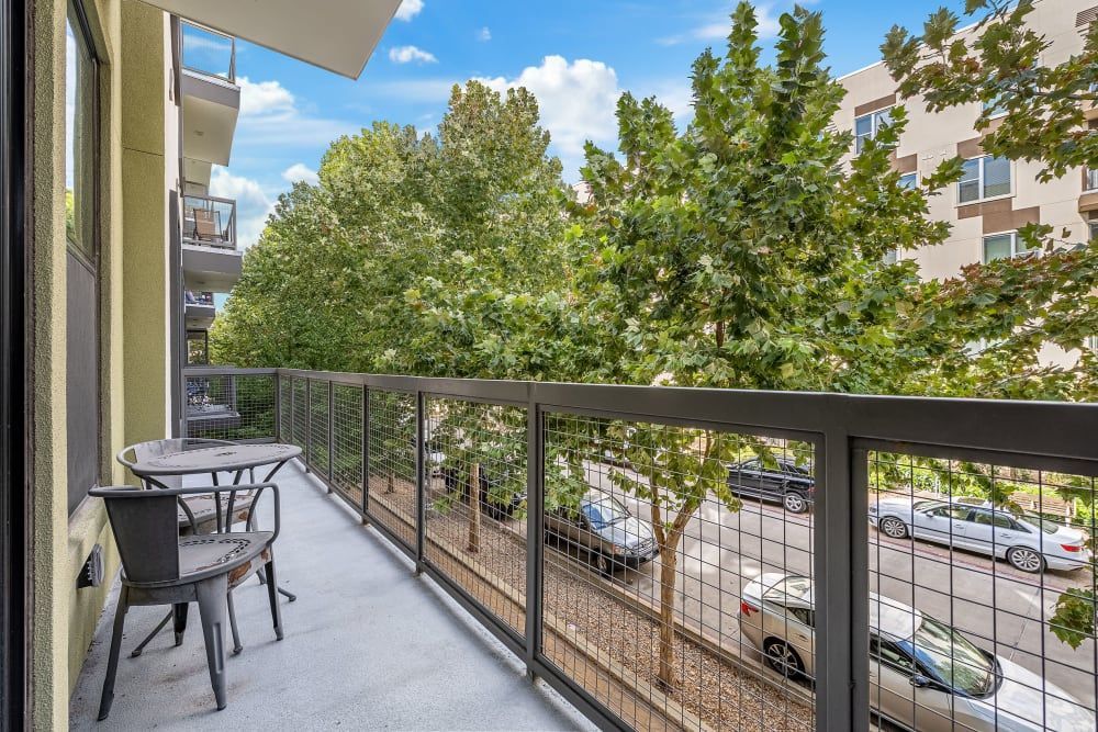 View from an apartment balcony showing a small patio table and chairs set against the backdrop of lush green trees and a parking lot with cars below. The balcony features a metal railing and overlooks a residential area with multi-story buildings at Marq Uptown in Austin, TX.