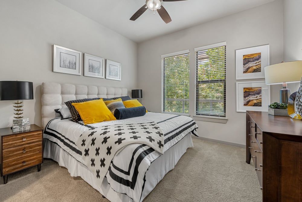 A modern bedroom featuring a large bed with a tufted headboard adorned with yellow and black pillows on top of patterned bedding. To the left, a wooden bedside table hosts a stylish lamp, while to the right, a sleek dresser stands beside a second lamp. Above the bed are framed abstract line art pieces, and on the adjacent wall, colorful landscape paintings hang above the windows draped with greenery outside. The room is lit with a ceiling fan and natural light, creating an inviting and cozy atmosphere at Marq Uptown in Austin, TX.