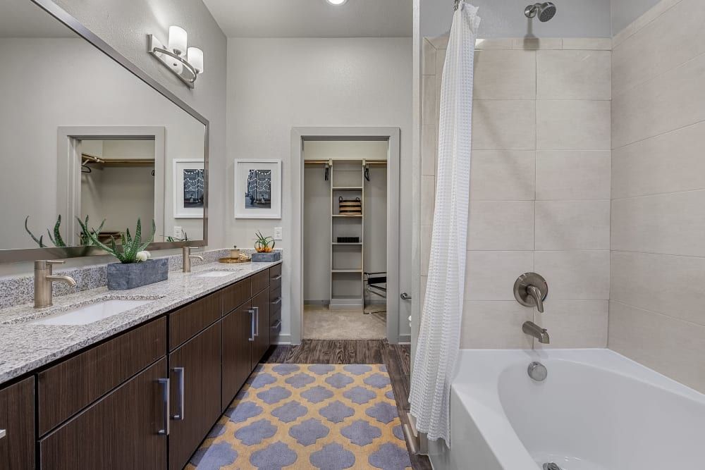 Modern bathroom interior with double vanity sink, large mirrors, and a walk-in closet in the background. A bathtub with a white curtain is on the right, complemented by beige tiled walls and patterned floor mat at Marq Uptown in Austin, TX.