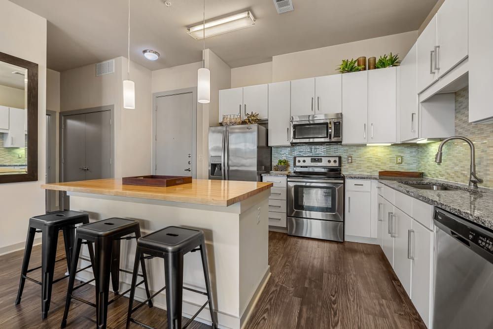 Modern kitchen interior with white cabinetry, stainless steel appliances, and a wooden countertop island with black stools. The kitchen features a tiled backsplash in shades of yellow and green, with pendant lights hanging above the island. The floor is dark wood, and there is a large mirror on the left wall at Marq Uptown in Austin, TX.