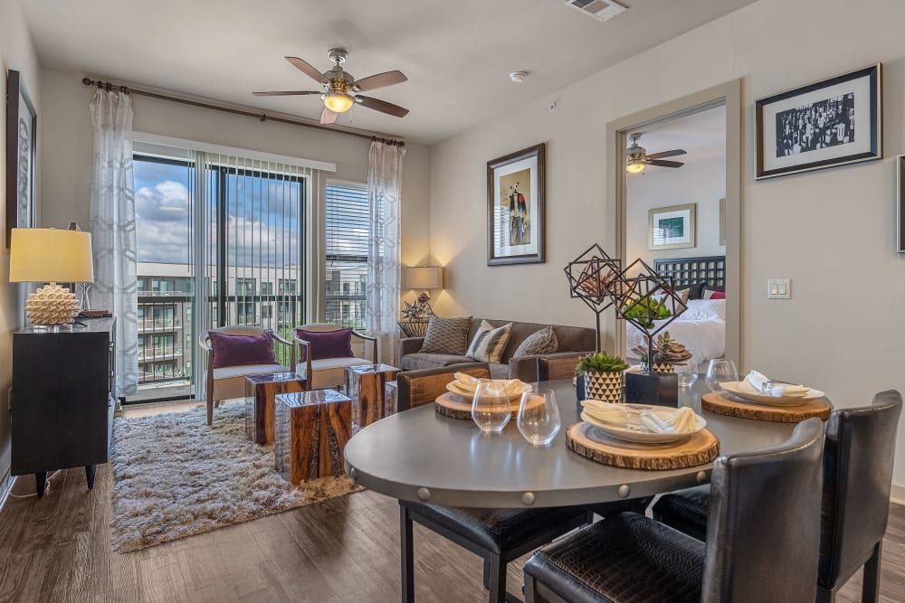 Contemporary living room with natural light coming through glass balcony doors, featuring a gray sofa, purple accent chairs, a fluffy area rug, and a dining area with a round table set for four at Marq Uptown in Austin, TX.