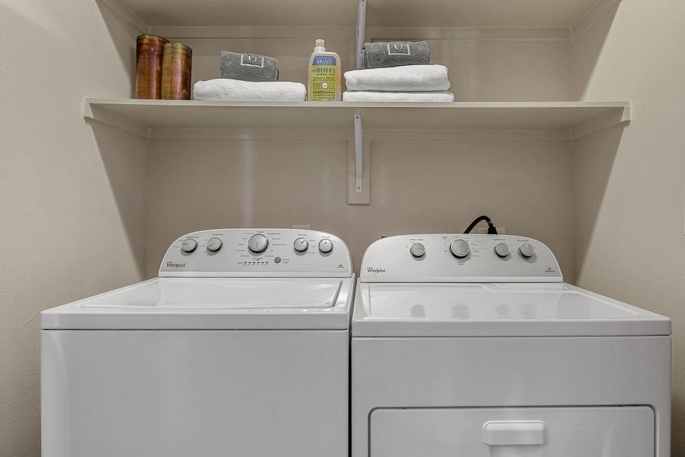 A modern laundry room with a white Whirlpool washing machine and dryer set. Above the appliances is a shelf stocked with folded towels, wooden containers, and a bottle of Mrs. Meyer's laundry detergent at Marq Uptown in Austin, TX.