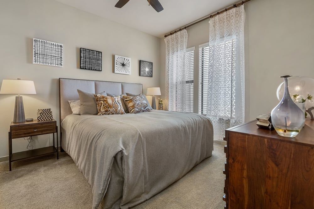 A well-lit, cozy bedroom with a neatly made bed featuring a tufted headboard and grey bedding with decorative pillows. A wooden bedside table on the left holds a lamp with a zigzag patterned base and a matching nightstand on the right has a clear vase with a unique design. Above the bed, a variety of black and white wall art creates a modern look, and light flows through white patterned curtains on the window. The room is completed with a beige carpet and a ceiling fan above at Marq Uptown in Austin, TX.