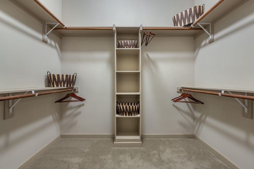 An empty walk-in closet with neutral-colored walls and carpet flooring, featuring wooden shelves, hanging rods and multiple wooden hangers. Some of the shelves have decorative baskets, and there is ample storage space on either side of a central, taller cabinet section at Marq Uptown in Austin, TX.