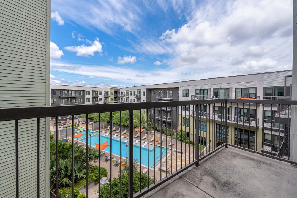 View from a balcony showcasing an apartment complex with modern grey buildings surrounding a communal pool area with lounge chairs and palm trees. The sky is partly cloudy at Marq Uptown in Austin, TX.