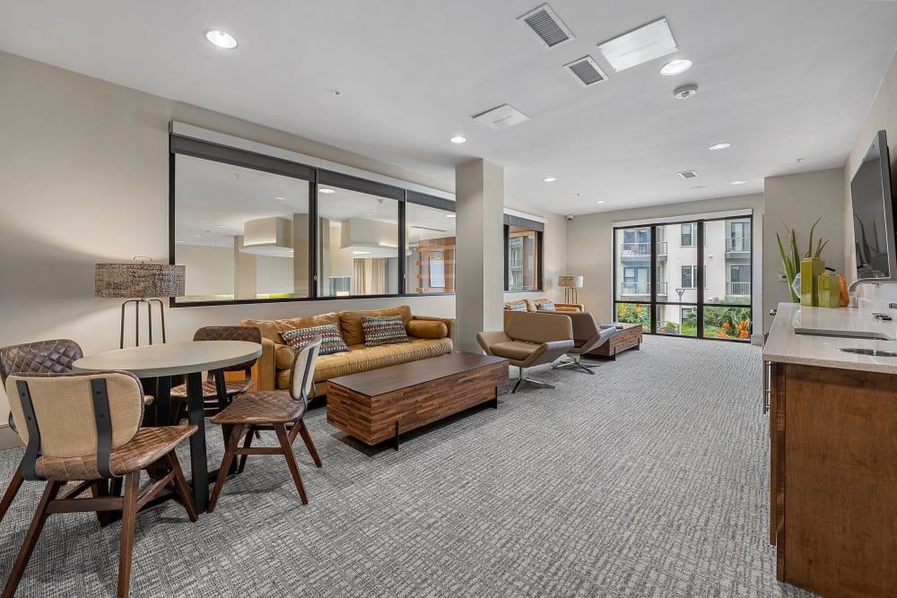 Modern and well-lit communal lounge area with comfortable seating including sofas and chairs, and a dining table. The room features large windows providing a view of the exterior, a flat-screen TV, and a kitchenette with bar counter. The decor is contemporary with a neutral color palette, and the space is carpeted at Marq Uptown in Austin, TX.
