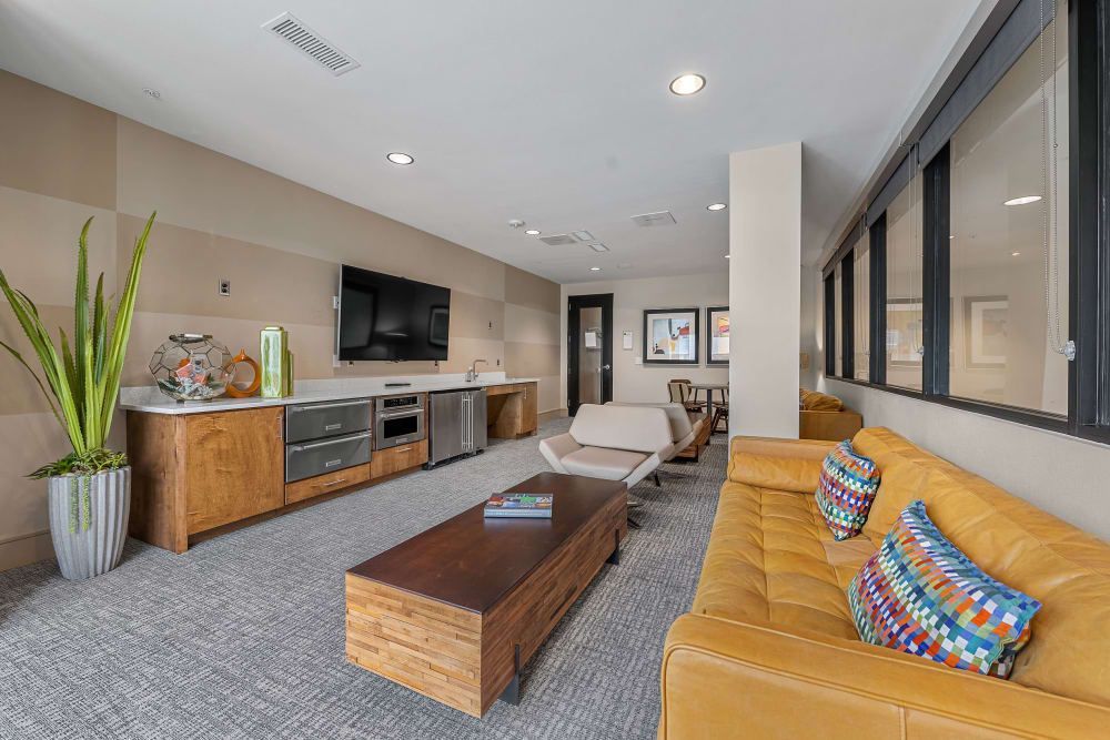 Modern living area with a spacious layout featuring a mustard-colored sectional sofa adorned with multicolored pillows, a stylish wooden coffee table, and a kitchenette with stainless steel appliances against the back wall. Large windows let in natural light, complementing the warm neutral tones of the room at Marq Uptown in Austin, TX.