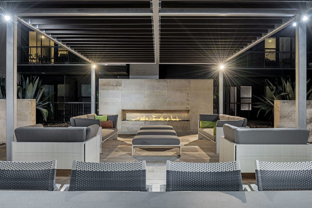 Modern outdoor patio area with comfortable seating and a long gas fireplace. The space features contemporary sofas and armchairs with gray cushions in front of a rectangular fire pit. Architectural elements include a dark pergola with illuminated ceiling, large concrete tiles on the floor, and structural columns. Ambient lighting creates a warm and inviting atmosphere. Lush plants add a touch of greenery to the sophisticated, neutral color palette of the design at Marq Uptown in Austin, TX.