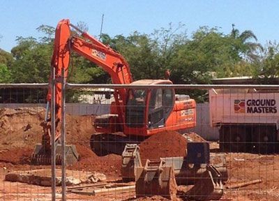 A large orange excavator is working on a construction site behind a fence.