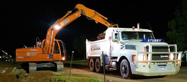 An excavator is loading dirt into a dump truck at night.