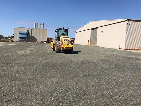A yellow roller is driving in a parking lot in front of a building.