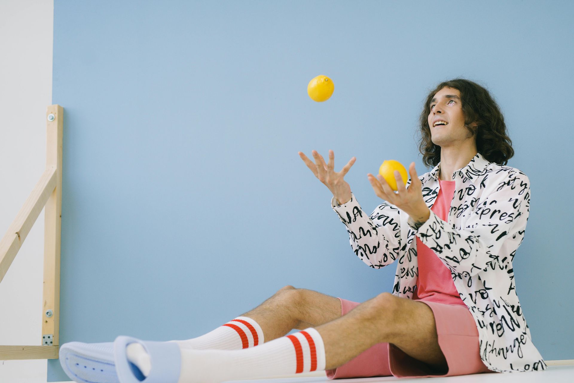 Man juggling lemons, seated against a blue wall. Wearing pink, white socks, light blue sandals, and a patterned jacket.