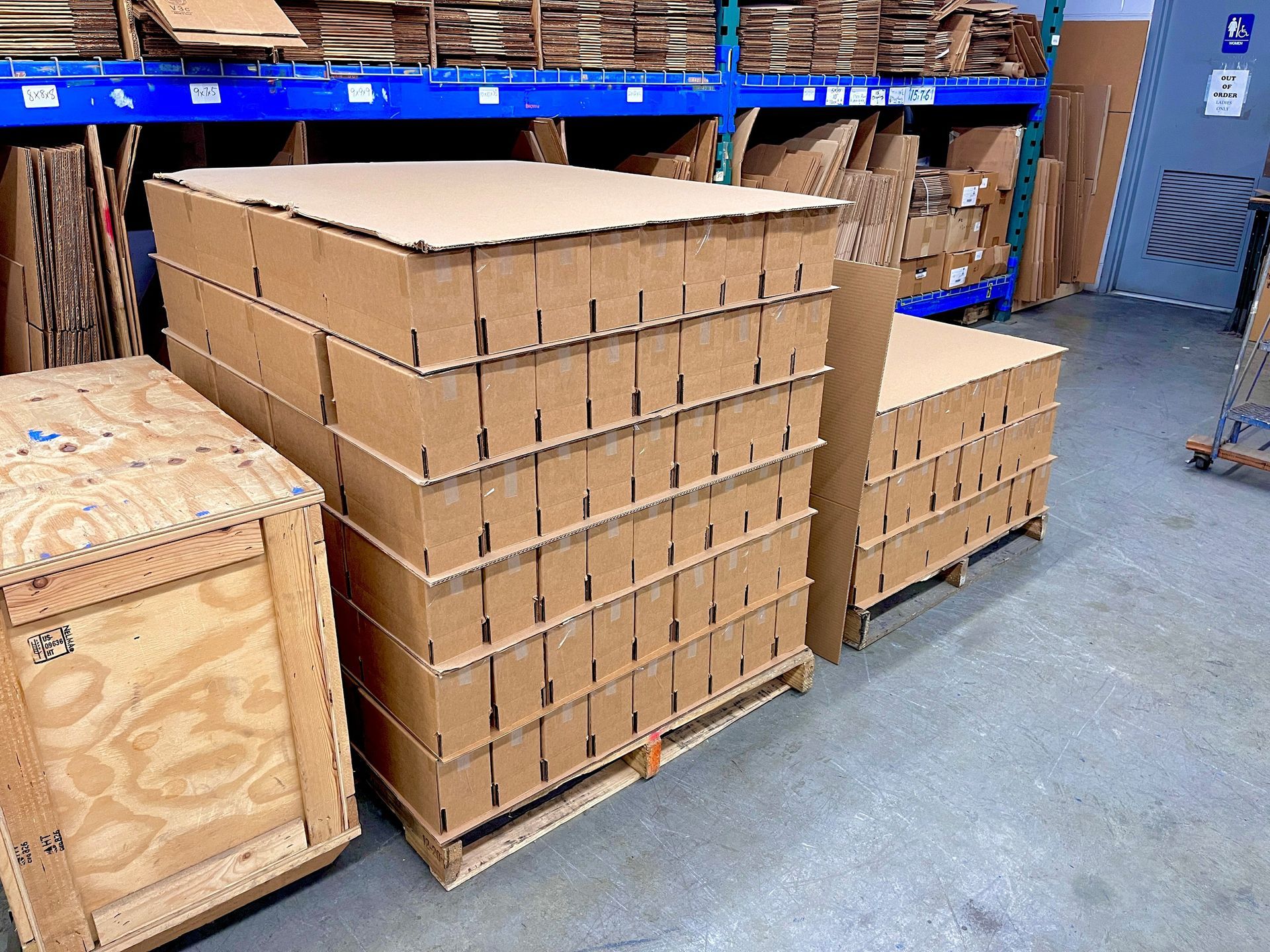 A pallet of many handmade, weather-resistant corrugated cardboard boxes. To its side is a wooden crate.