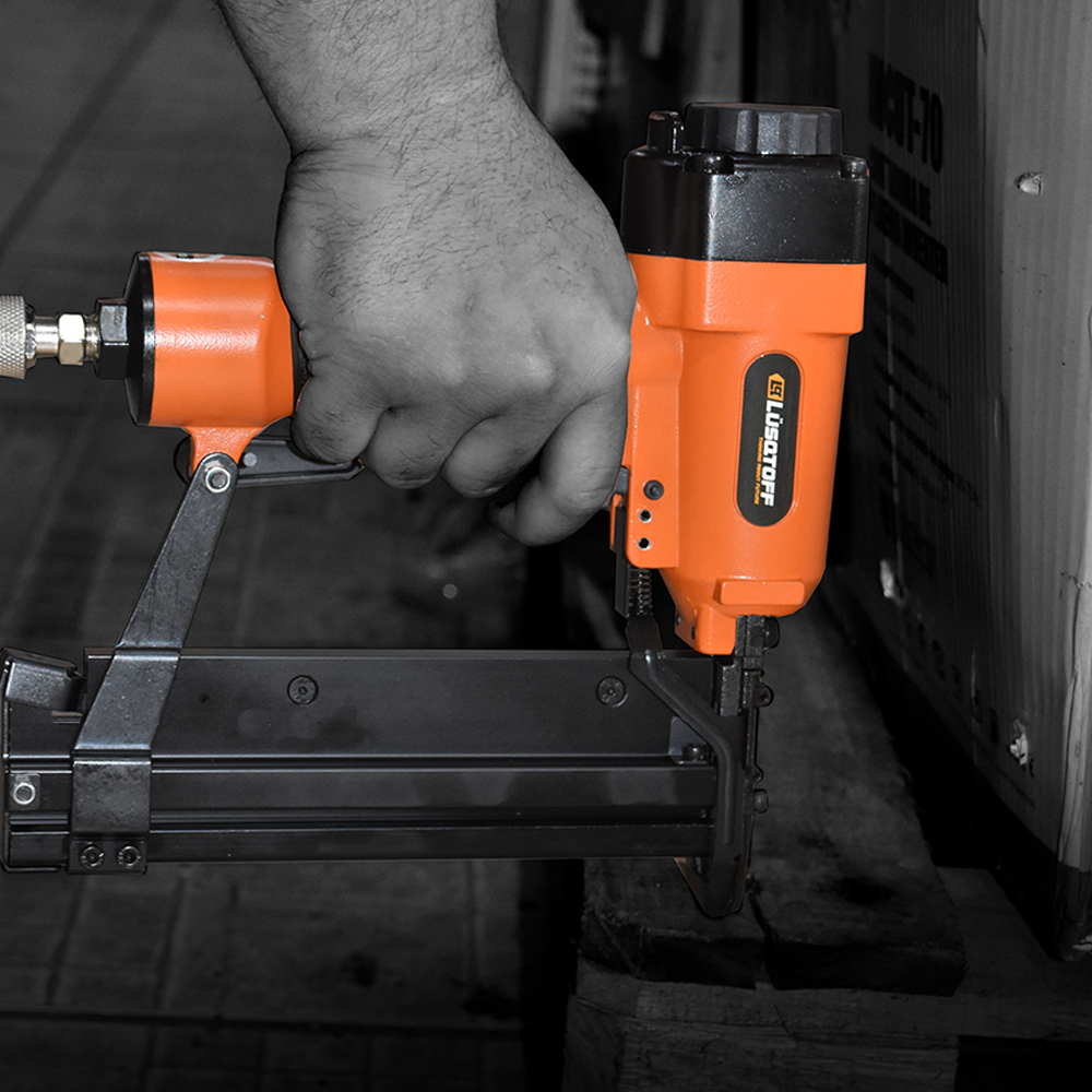 Persona que utiliza una pistola de clavos neumática naranja sobre una paleta de madera; fondo en escala de grises.