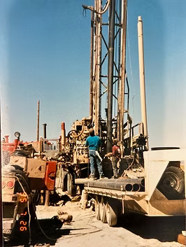 Two workers on a drilling rig in a desert setting. Rig is mounted on a truck bed. Clear blue sky.