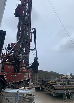 Person operating a red drilling rig outdoors on a cloudy day. Pipes are stacked nearby.