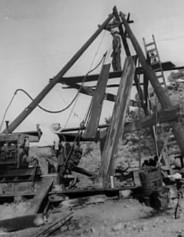 Workers operating a wooden well drilling rig outdoors. One stands on the platform, others operate machinery.