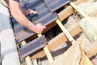 A person installing dark roof tiles onto a wooden roof frame with insulation visible nearby.