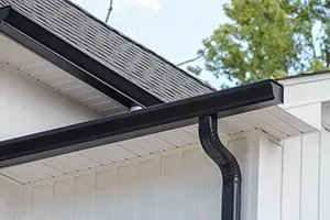 A black gutter and downspout system installed on the white siding of a home with a shingled roof.