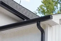 A black gutter and downspout system installed on the white siding of a home with a shingled roof.