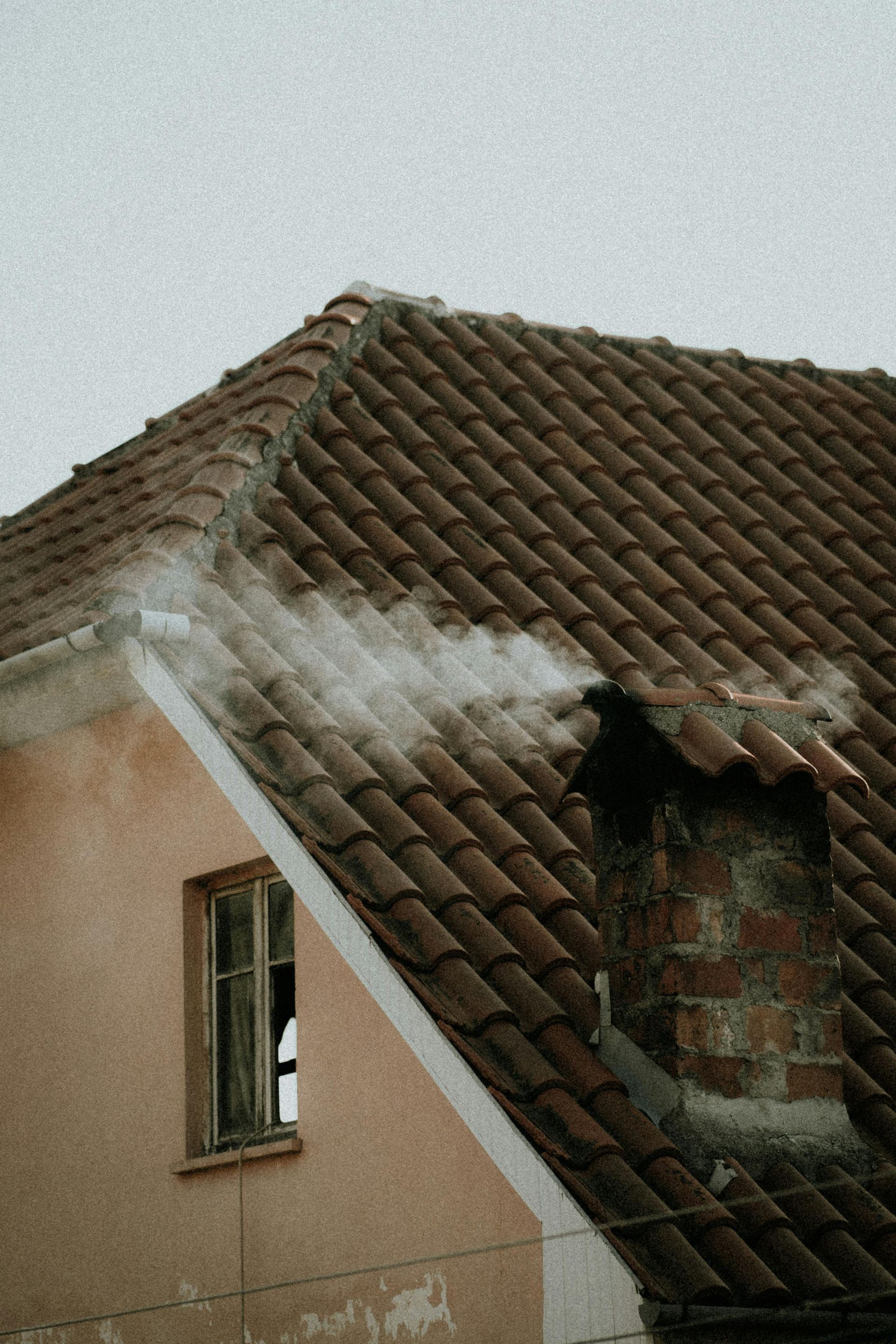A section of a peach-colored building with a brown tiled roof, featuring a broken window and a damaged roof section.